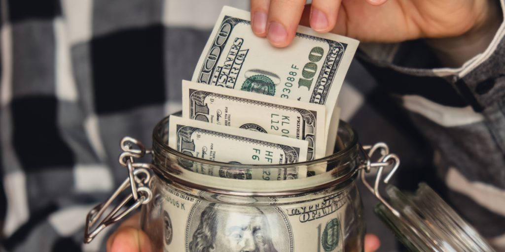 Hand placing US dollar bills into a glass jar, representing saving money and budgeting.
