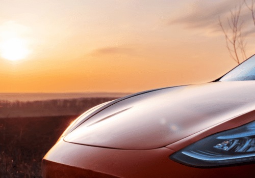 Close-up of a red car hood and headlight at sunset, with warm light reflecting across a scenic view.
