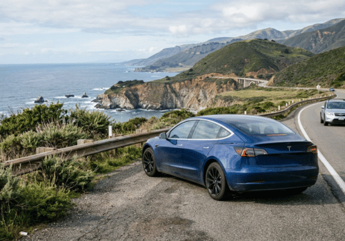 Blue Tesla parked along a scenic coastal highway with ocean views, cliffs, and a winding road. 