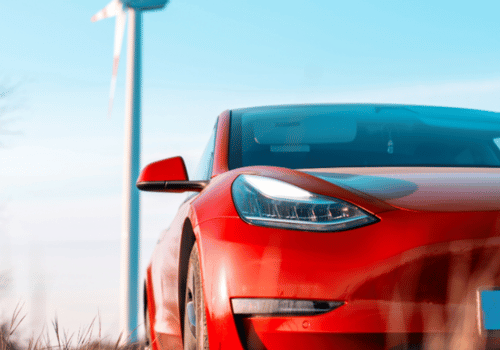 Red Tesla parked outdoors with a wind turbine in the background. 