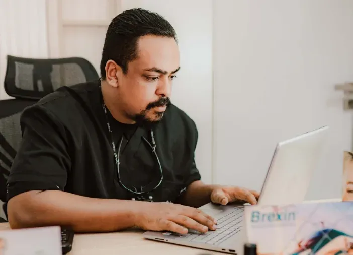Une personne vêtue d'une chemise noire tape sur un ordinateur portable à un bureau, dans un environnement de bureau.