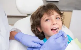 Un dentiste portant des gants bleus utilise un miroir buccal pour examiner un enfant installé sur un fauteuil dentaire.