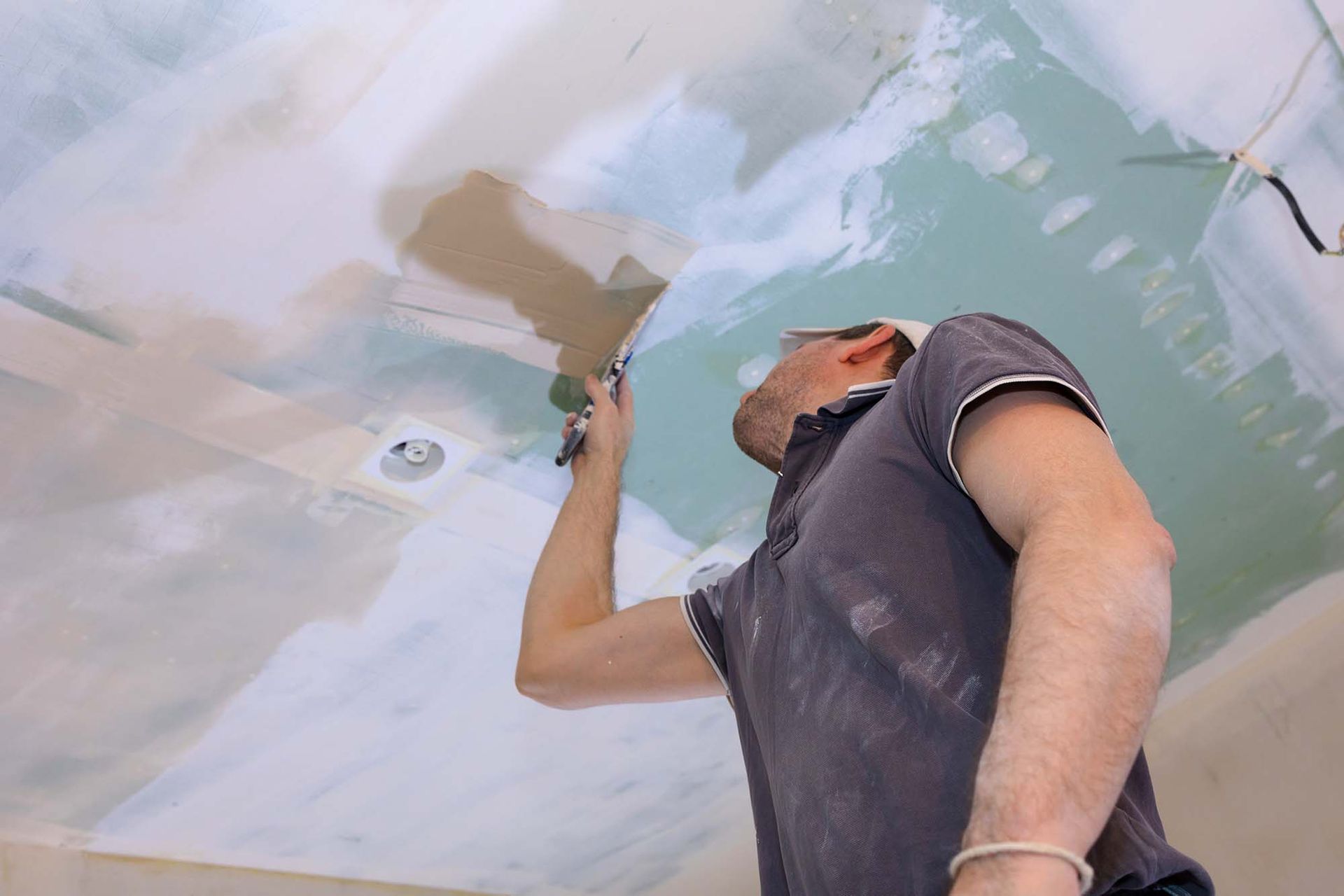Worker sanding a ceiling with a tool, wearing a dust-covered shirt in a room under renovation