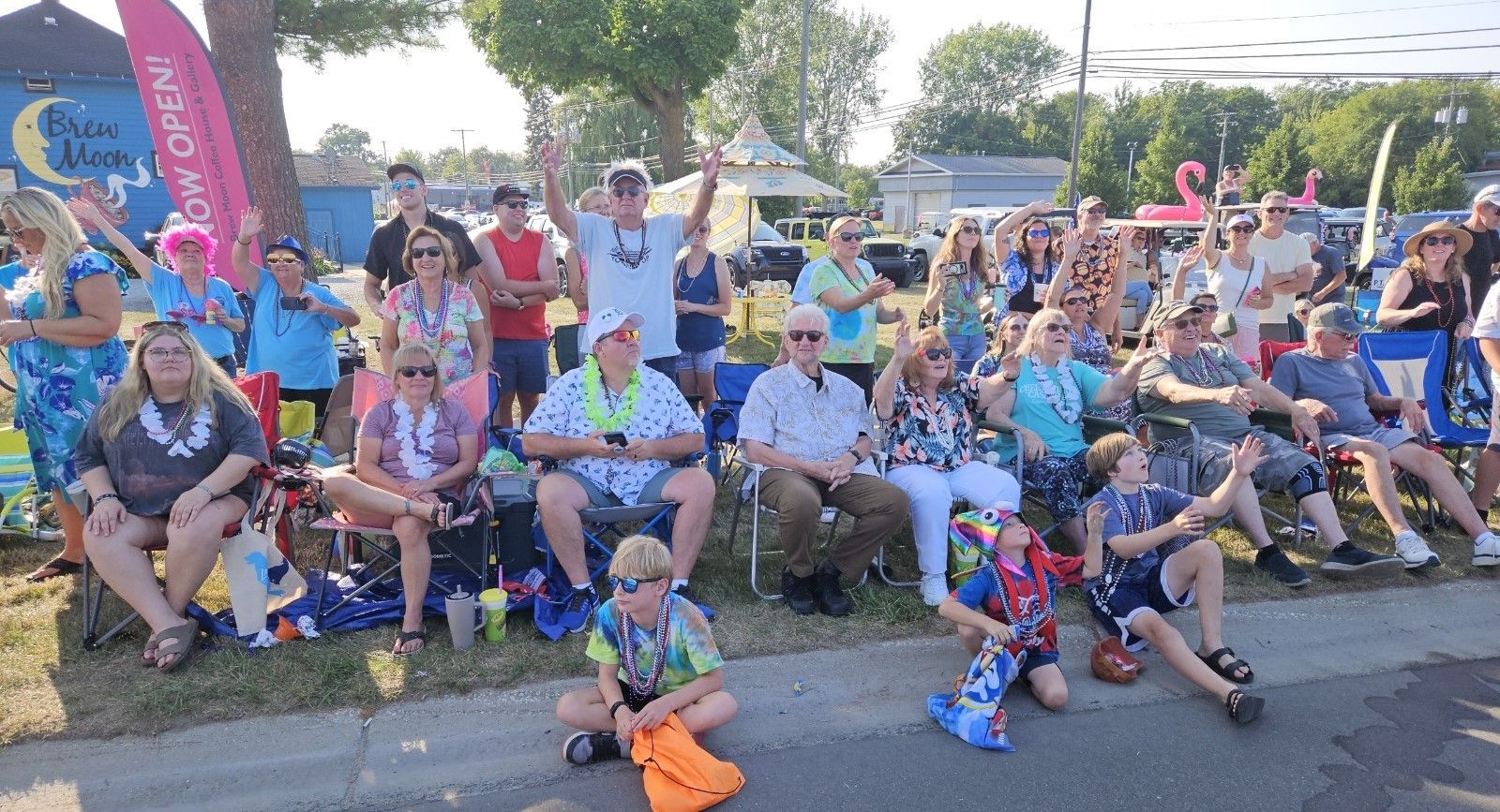 A large group of people are sitting on the side of the road watching a parade, the cheeseburger fest