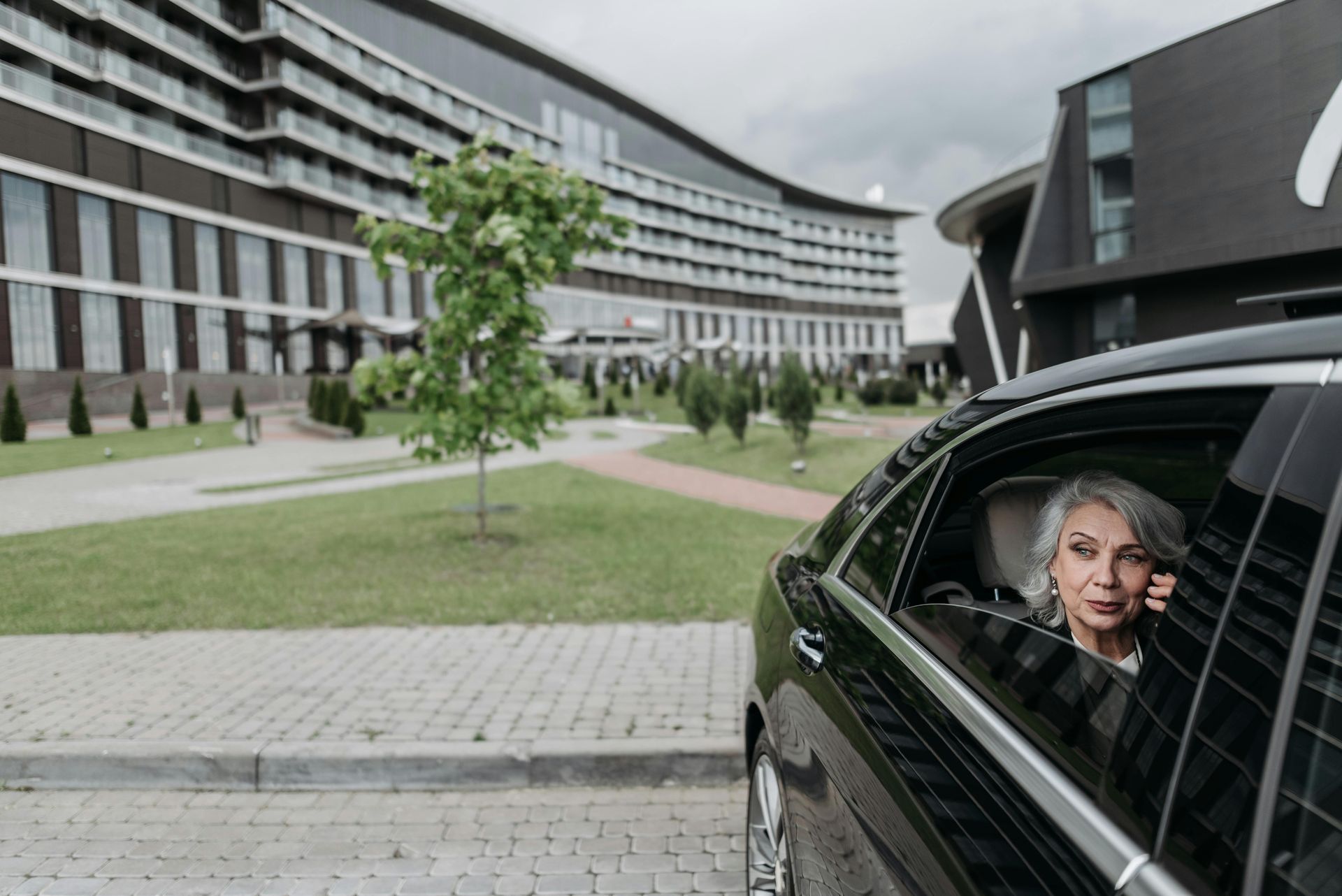 Woman in car looks toward a modern building. Gray hair, dark car, overcast day.