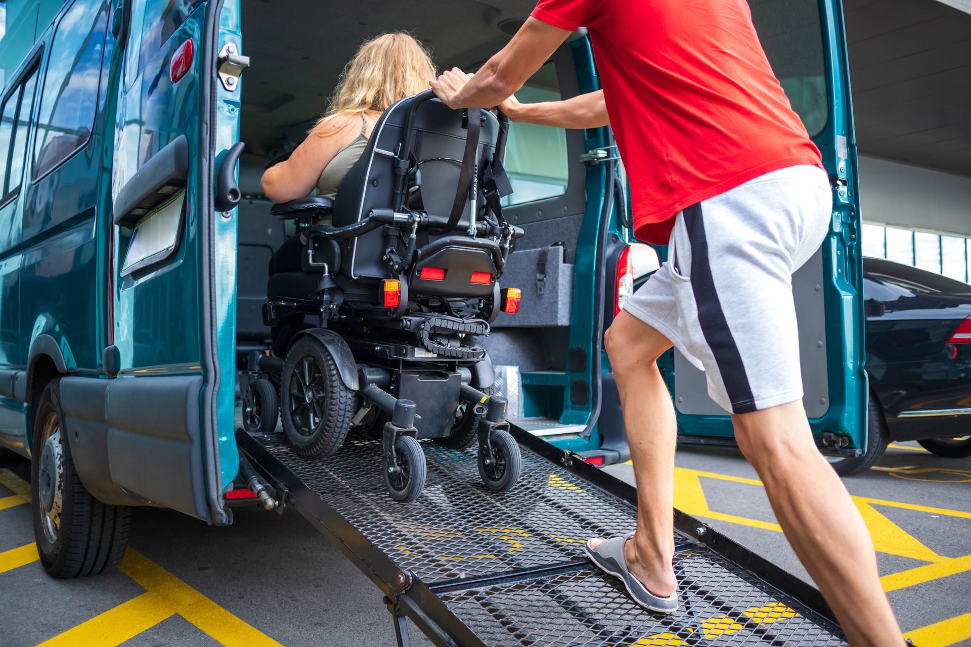 Man assisting woman in wheelchair up a ramp into a teal accessible van in a parking lot.