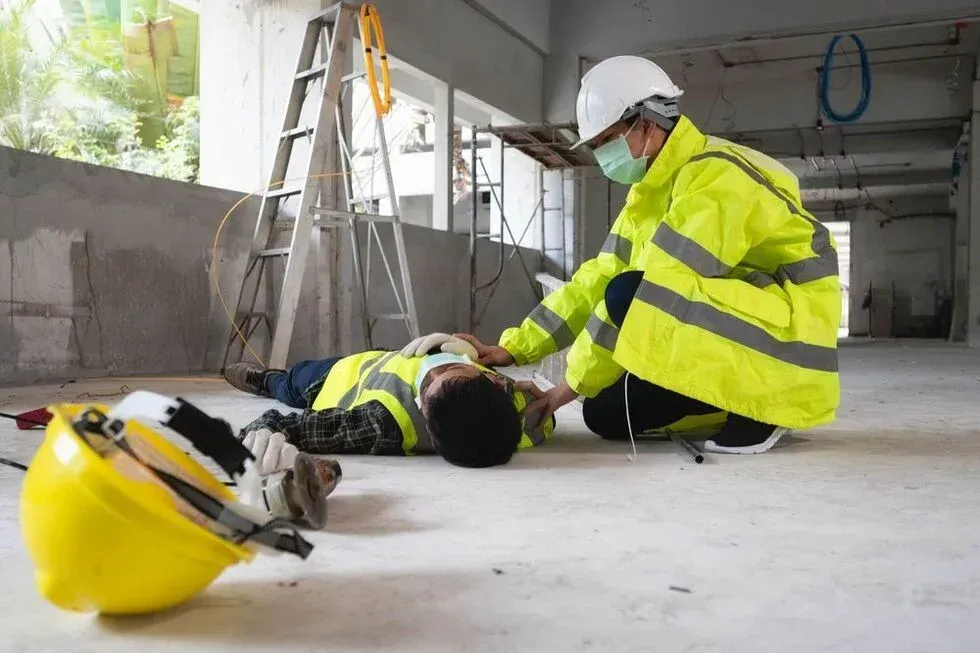 Construction worker collapsed on floor, being checked by a colleague wearing safety gear, inside building.