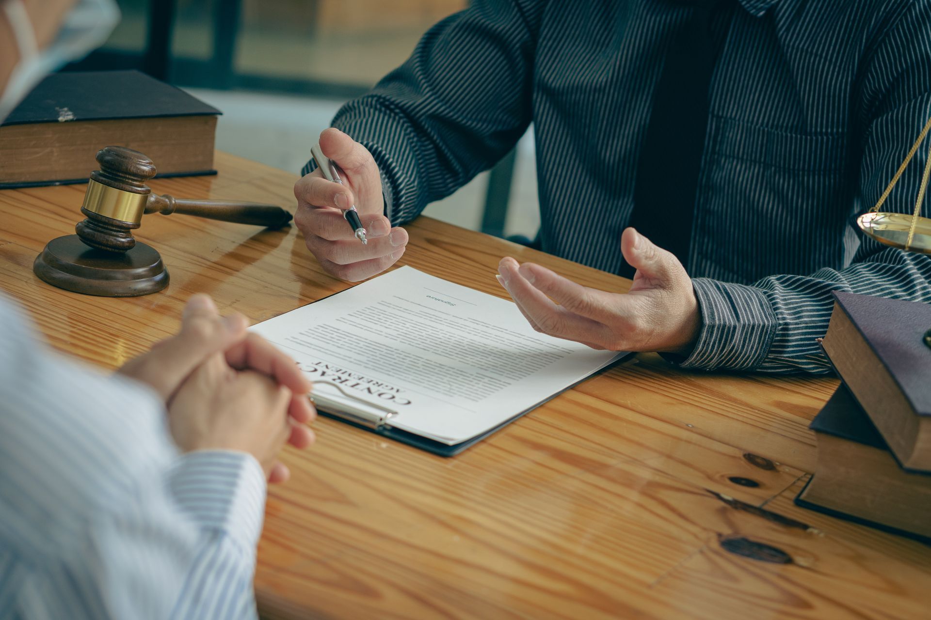 Two people at a desk, reviewing paperwork. One gestures with a pen, the other clasps their hands. Gavel and scales of justice are present.