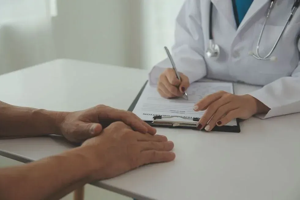 Doctor writing on a clipboard, hands clasped with a patient at a desk.