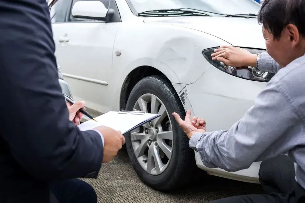 Two men inspecting damage on a white car; one holds a clipboard, the other points at the dented fender.