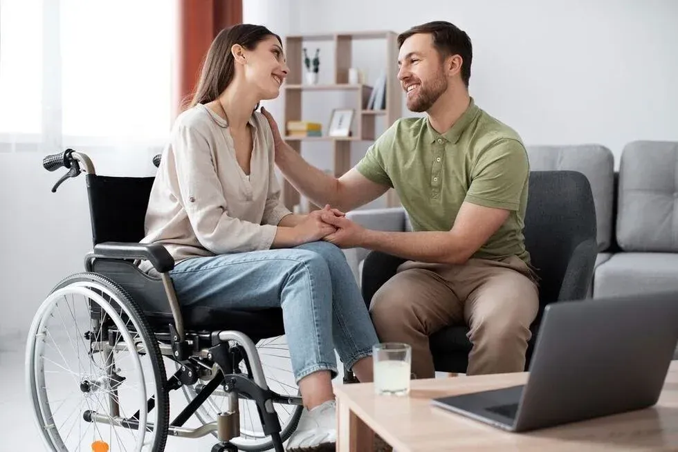 Woman in wheelchair and man hold hands, looking at each other. They smile, touching hands, in a living room with a laptop.