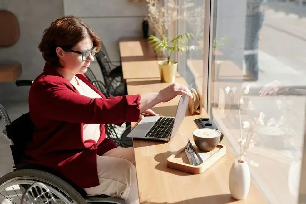 Woman in wheelchair, red blazer, working on laptop at a cafe table by a window; coffee cup.