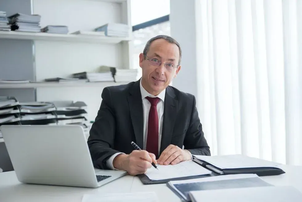 Man in suit at desk, writing, with laptop and documents. Office setting.