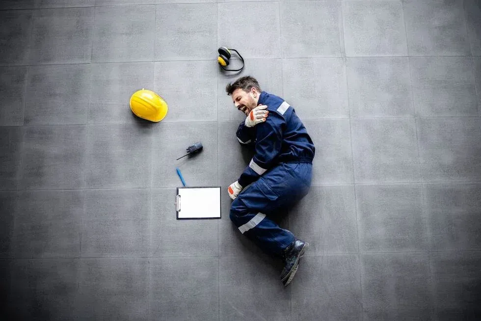 Worker in blue uniform lies on gray floor, clutching shoulder, with fallen helmet and clipboard.