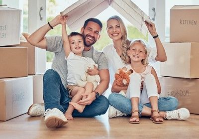 Family seated on the floor, surrounded by boxes, making a roof with cardboard, smiling.