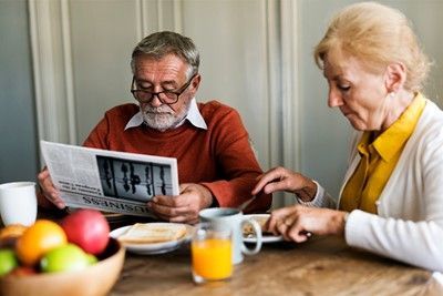 Older adult reads newspaper while another eats at a table with breakfast, coffee, and juice.