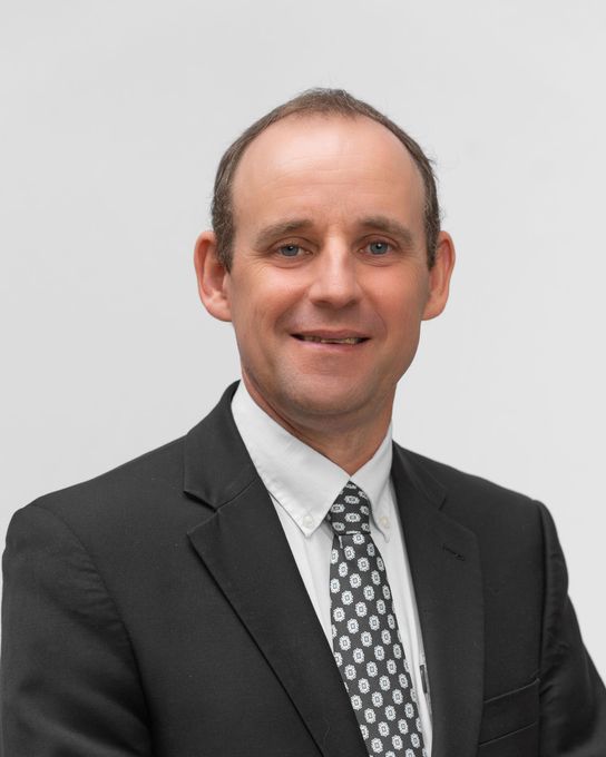 Man in black suit and patterned tie, smiling against a white background.