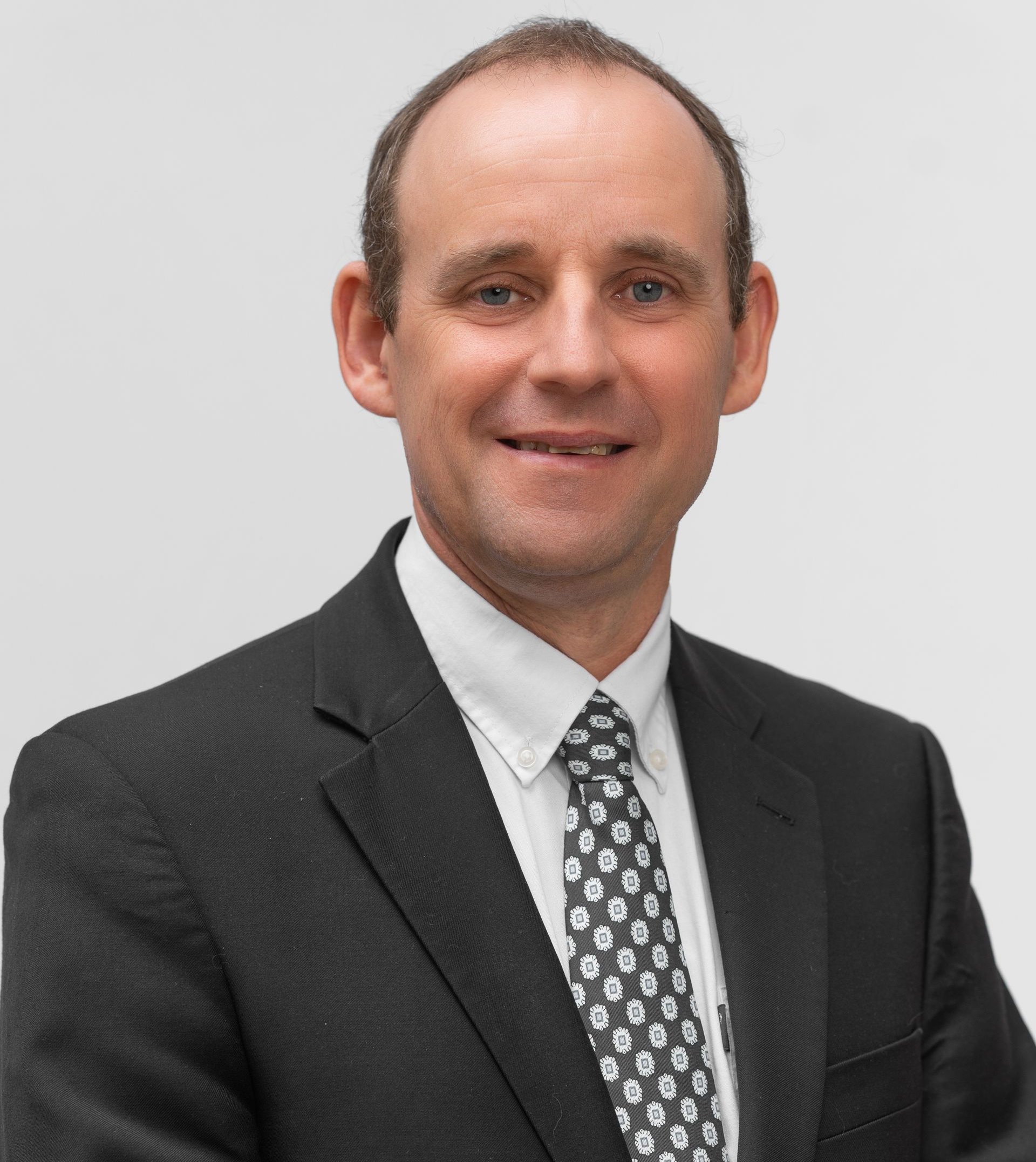 Man in a suit smiles at the camera, with a white collar and patterned tie against a white background.