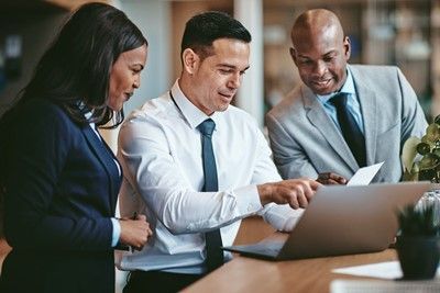 Three professionals reviewing a laptop screen. The central person points at the screen. Office setting.