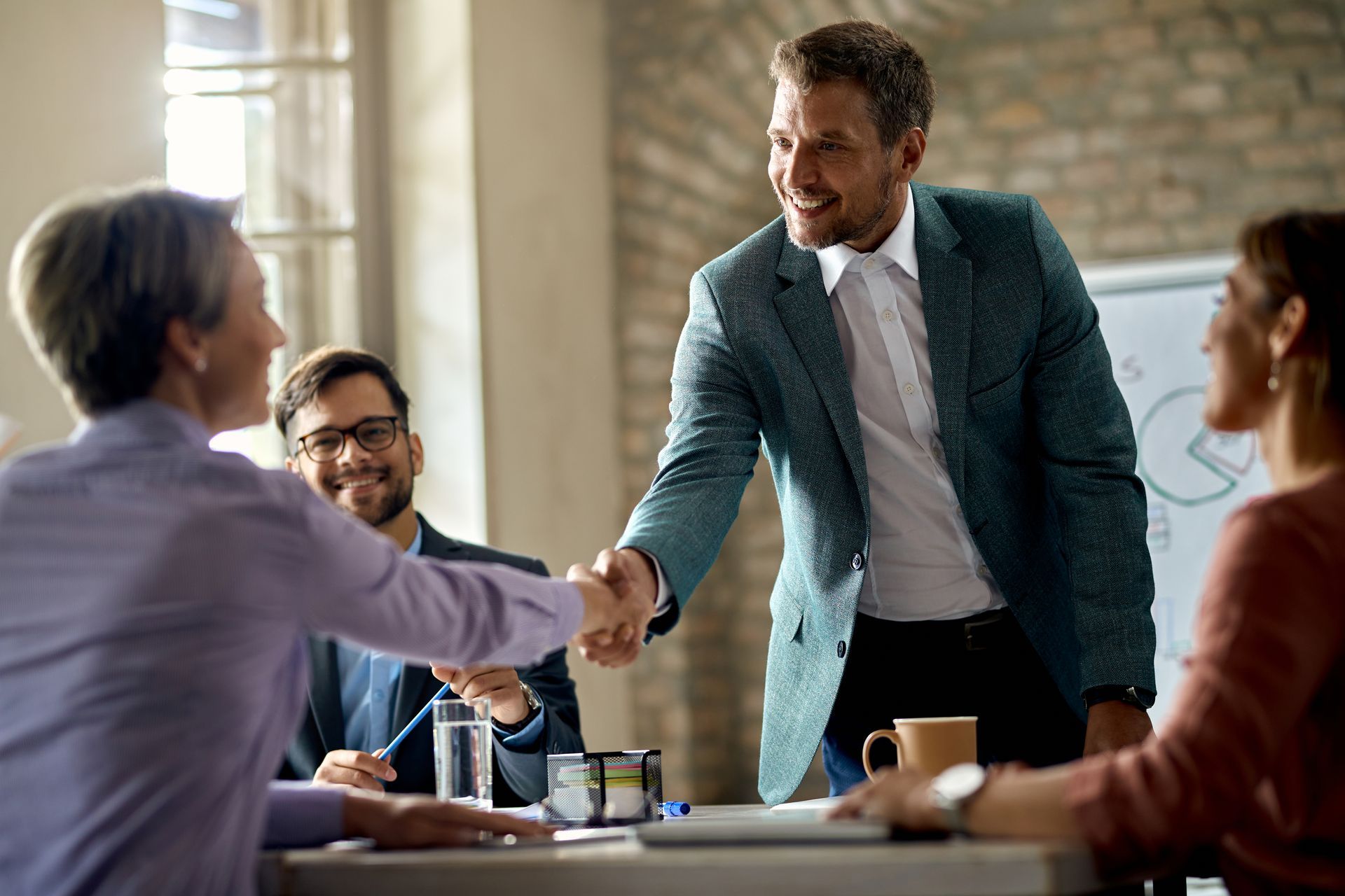 Businesspeople in an office, shaking hands and smiling. Other colleagues sit at a table.