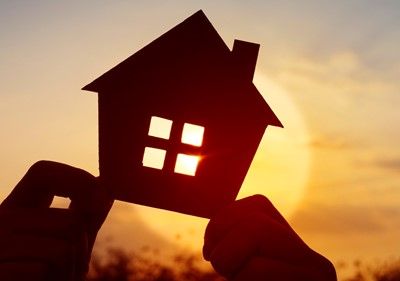 Hands holding a cutout house silhouette against a setting sun.