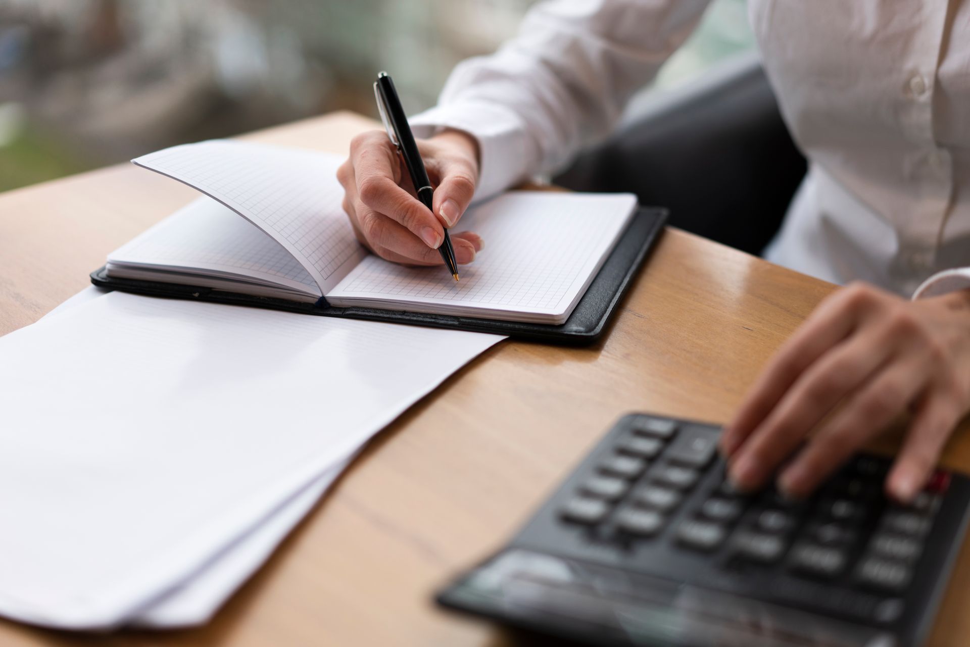 Person writing in a notebook, using a calculator, and working on documents at a desk.