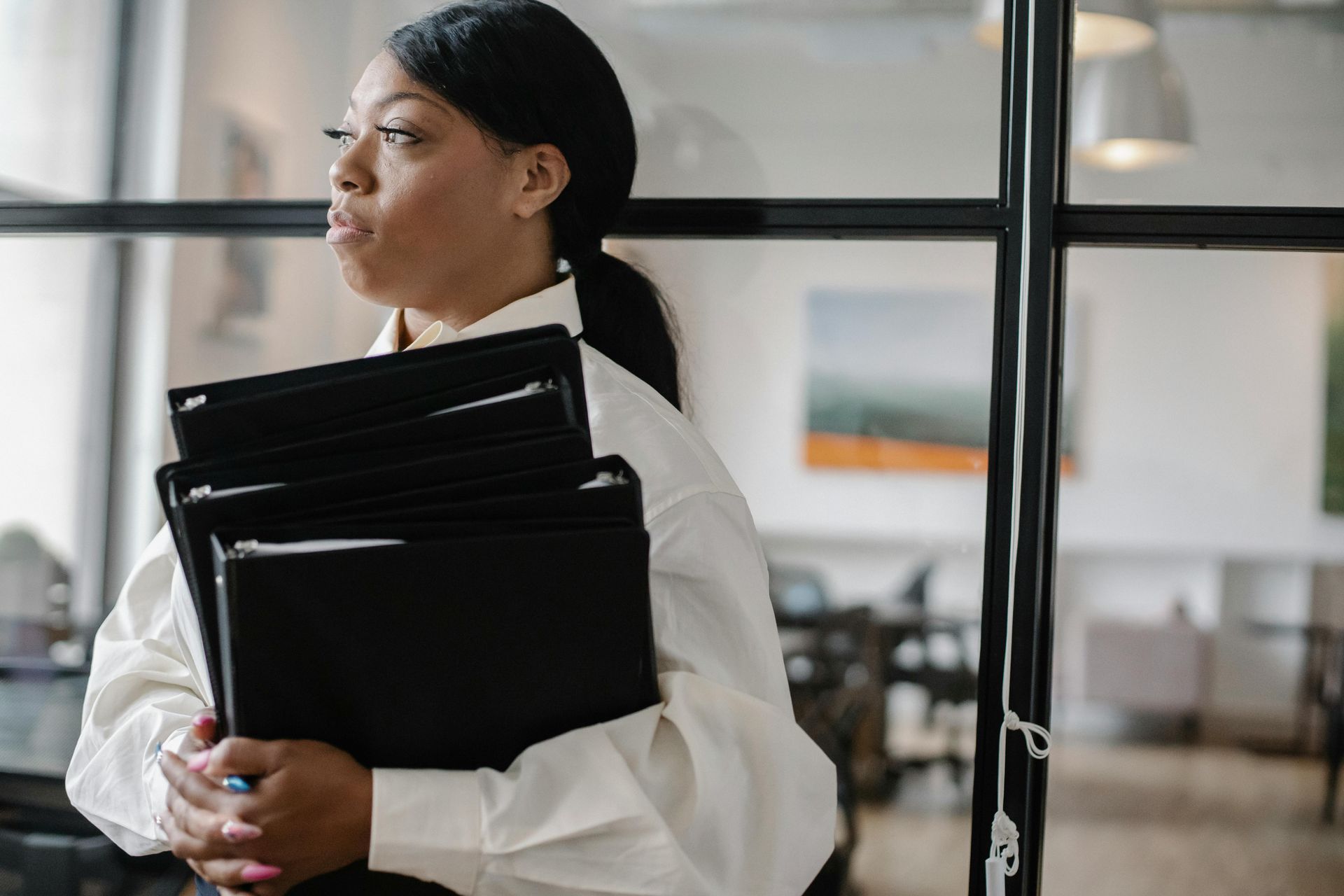 Woman in white shirt holds black folders, looking thoughtful near glass wall.