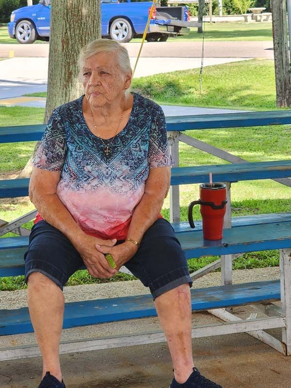 An elderly woman is sitting at a picnic table with a red cup on it.