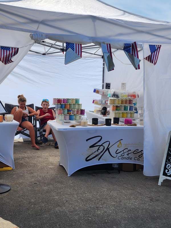 A woman sits at a table under a tent with a table cloth that says 3 kisses on it