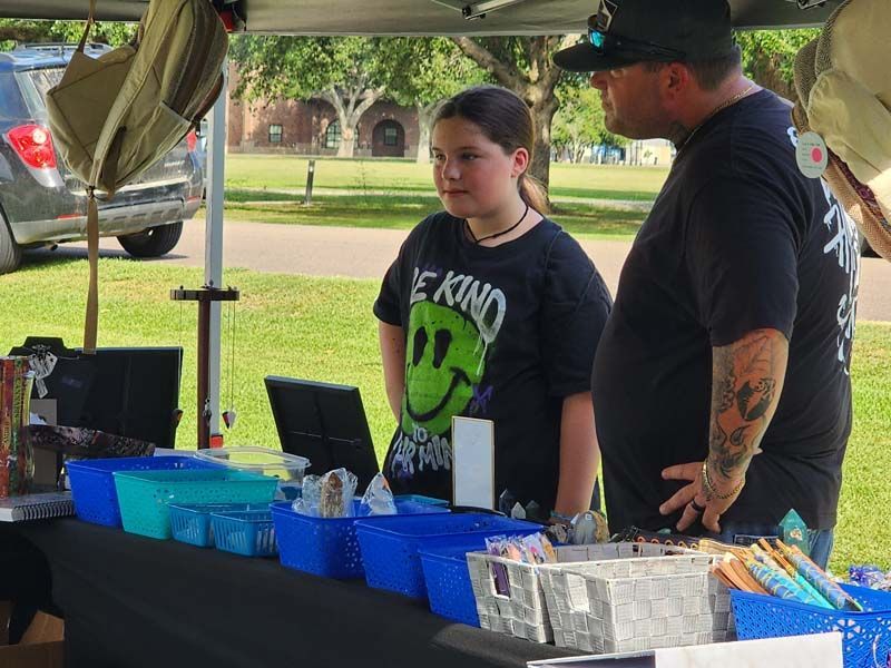 A man and a girl are standing in front of a table . the girl is wearing a shirt that says be kind.