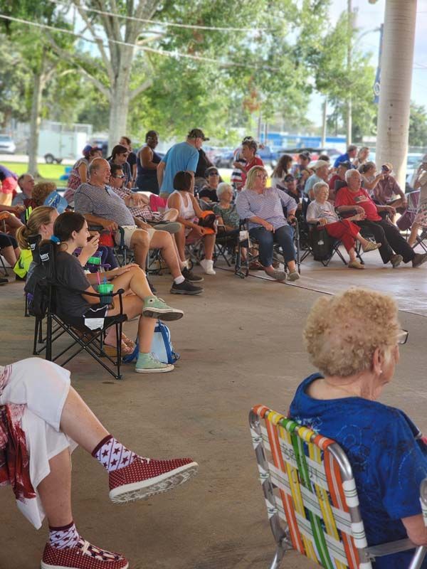 A group of people are sitting in folding chairs under a covered area.