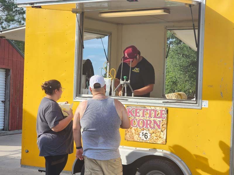 Two people are standing in front of a yellow food truck that sells kettle corn for $ 6
