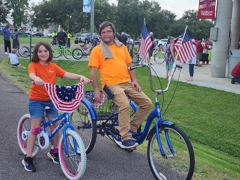A man and a girl are riding tandem bicycles decorated with american flags.