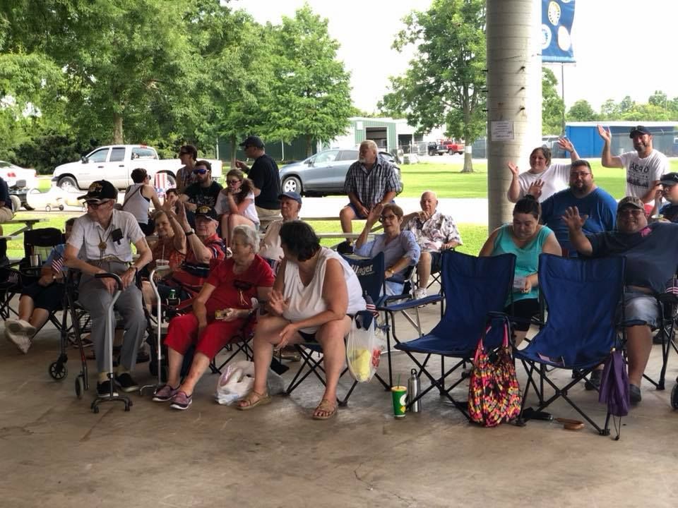 A group of people are sitting in folding chairs under a covered area.