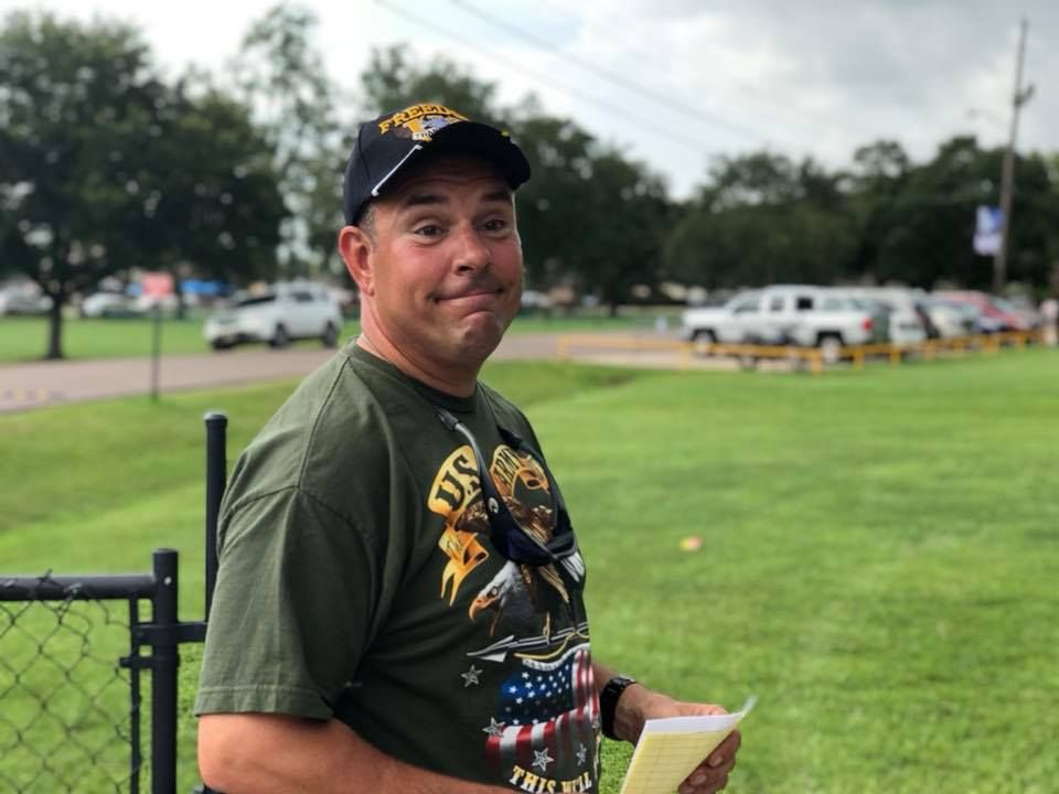 A man wearing a hat and a t-shirt is standing in a park holding a piece of paper.
