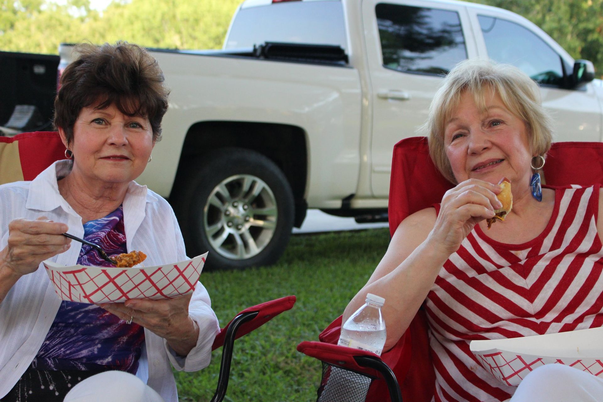 Two women are sitting in chairs eating food in front of a truck.
