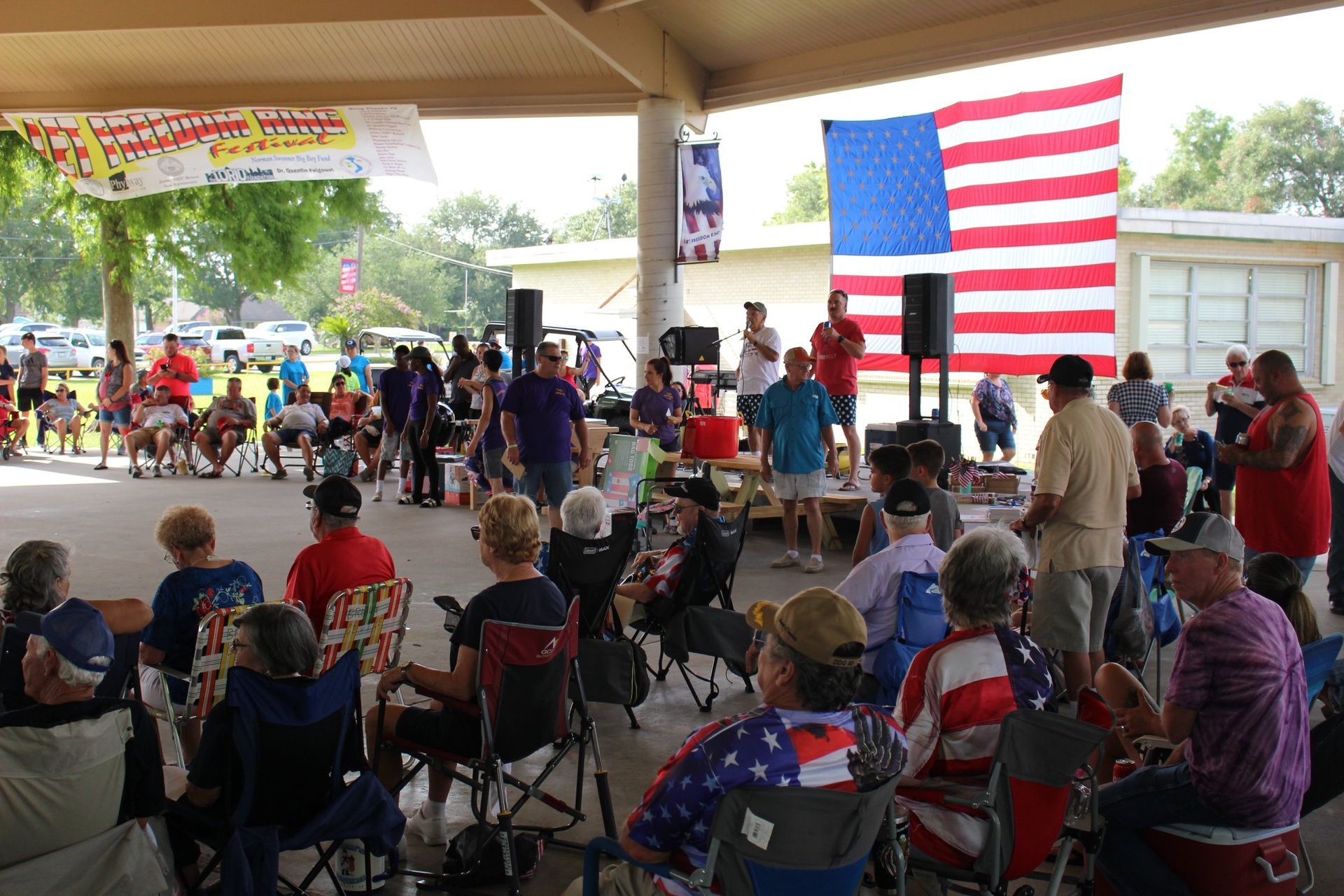 A group of people are sitting in chairs under a pavilion with an american flag in the background.
