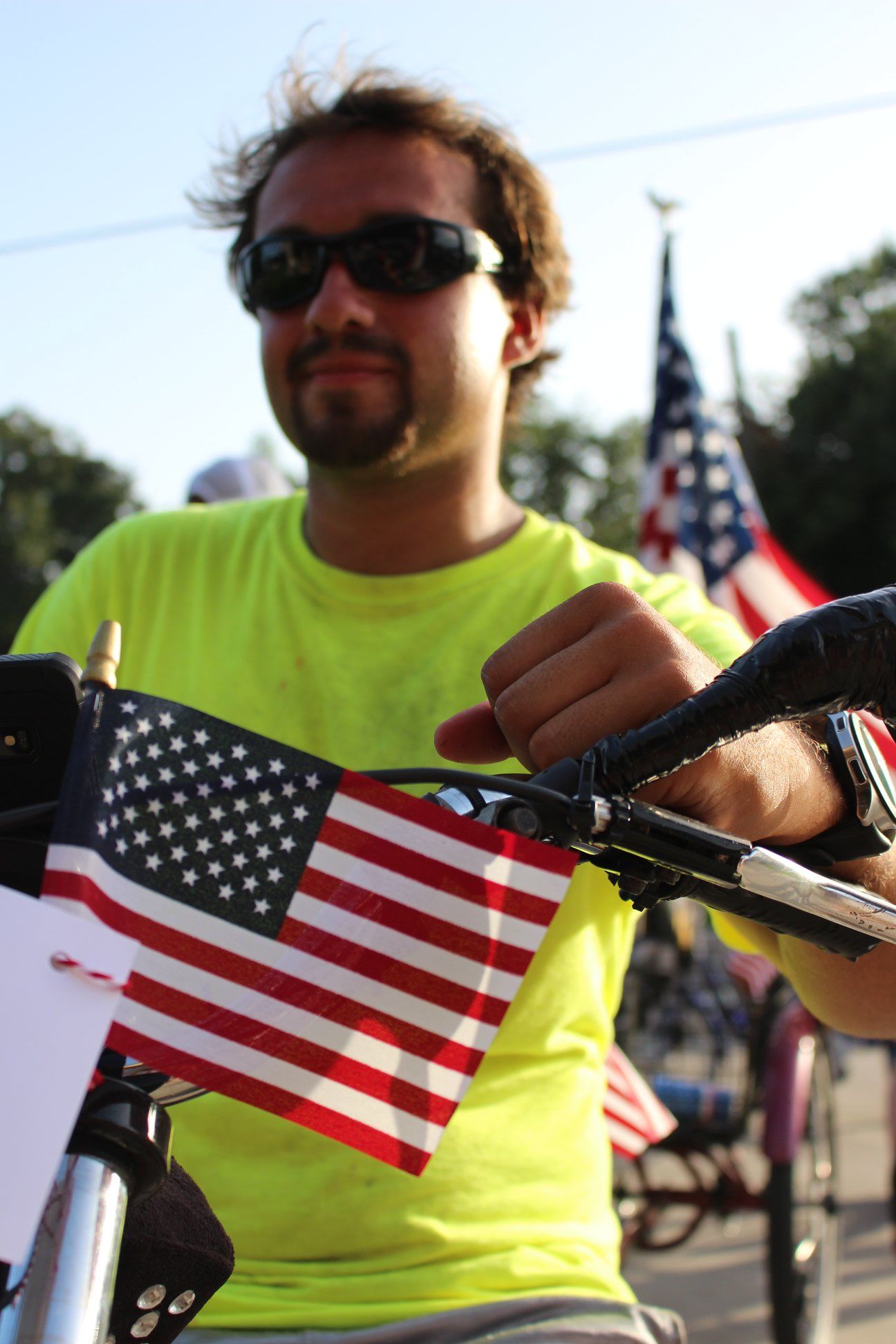 A man wearing sunglasses is riding a bike with an american flag attached to the handlebars
