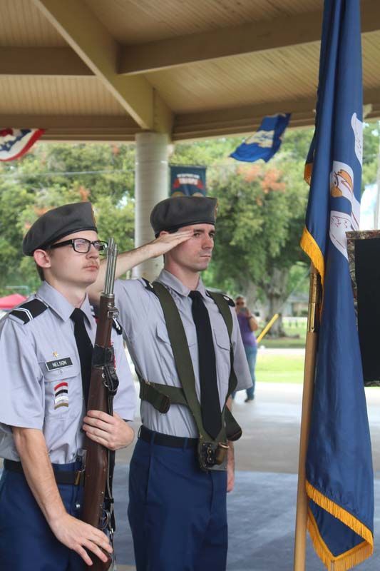 Two men in military uniforms salute in front of a flag