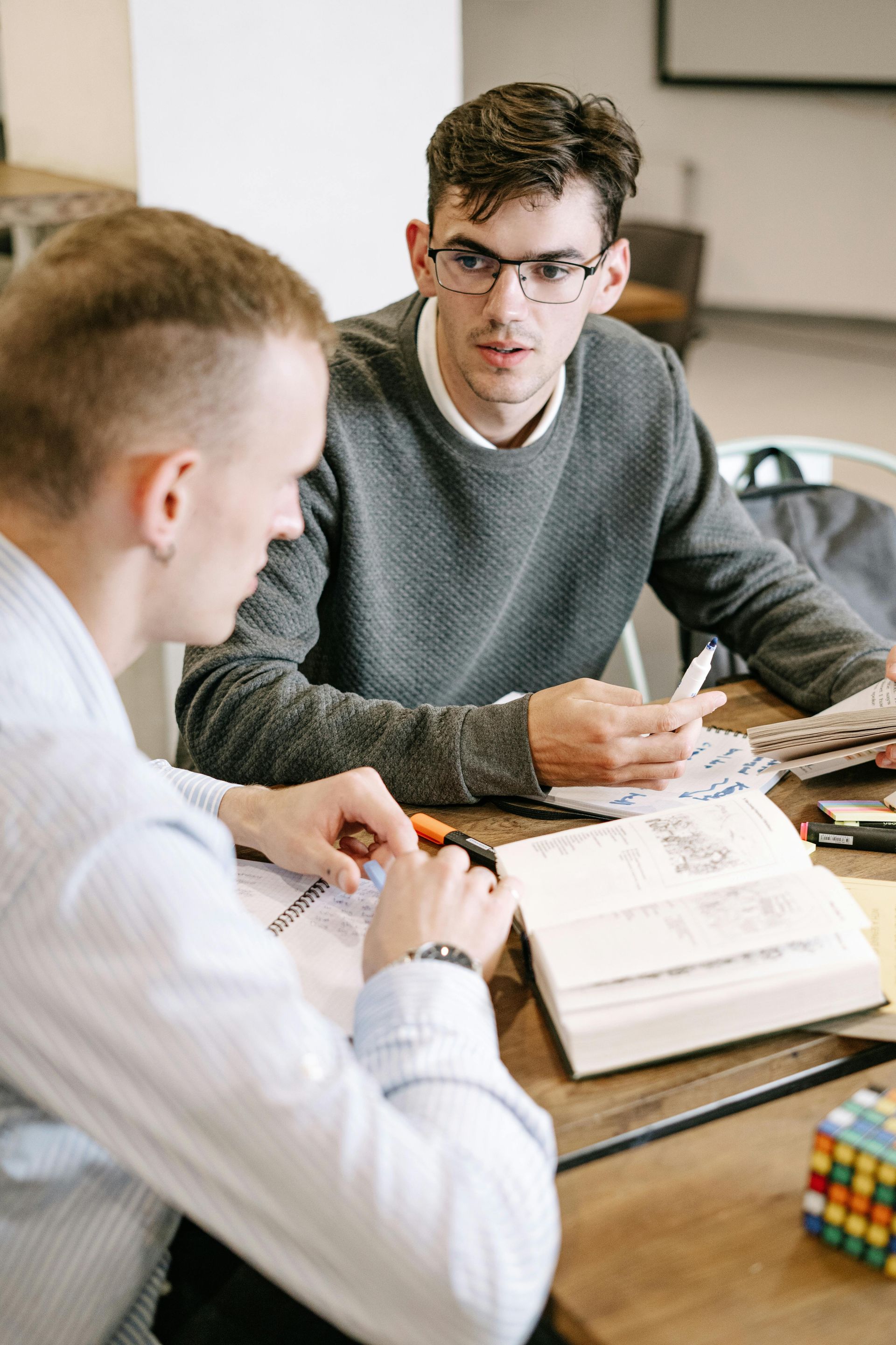 Two men are sitting at a table with a book and a rubik 's cube.