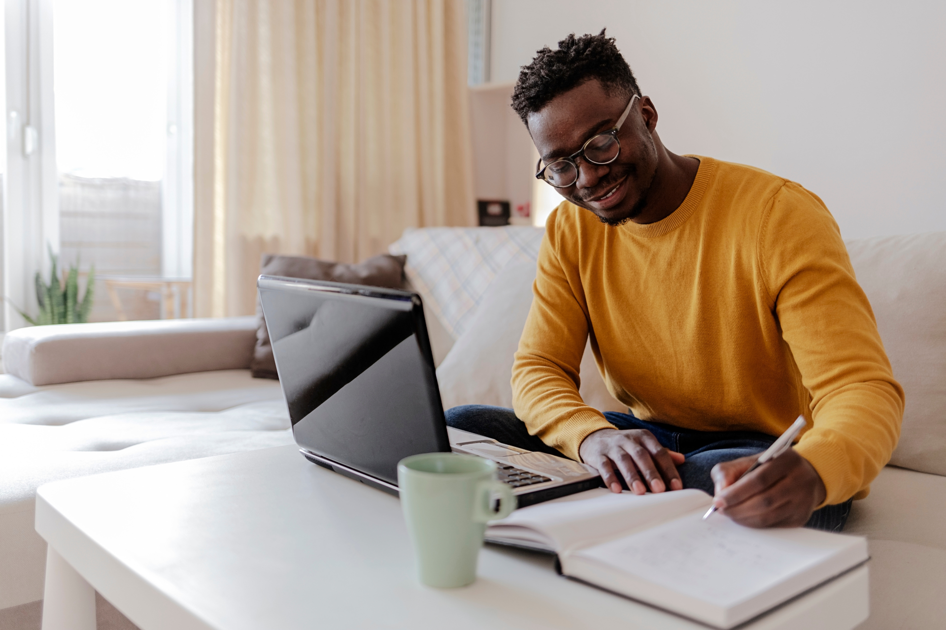A man is sitting on a couch using a laptop and writing in a notebook.