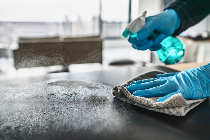 A person wearing blue gloves is cleaning a table with a cloth and spray bottle.