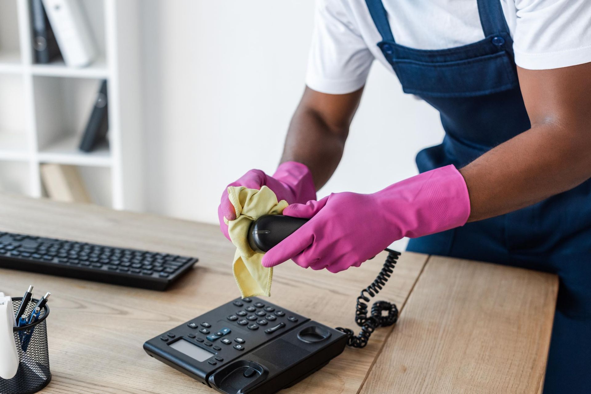A man in pink gloves is cleaning a telephone on a desk.