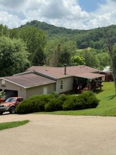 Beige house with a brown roof and green bushes in front, trees and mountains in the background.