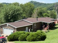 Brown house with attached garage, surrounded by trees and a grassy hill, with people standing near the front door.