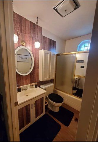 Small bathroom with wood-look accent wall, vanity, toilet, and shower/tub. Round mirror and cabinet.