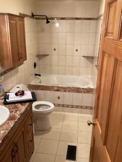 Bathroom with a toilet, vanity, and tub/shower. Beige tile, wooden cabinets, and a partially open wooden door.