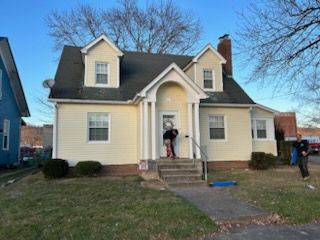 Yellow house with two dormers, steps leading to the door; people outside.