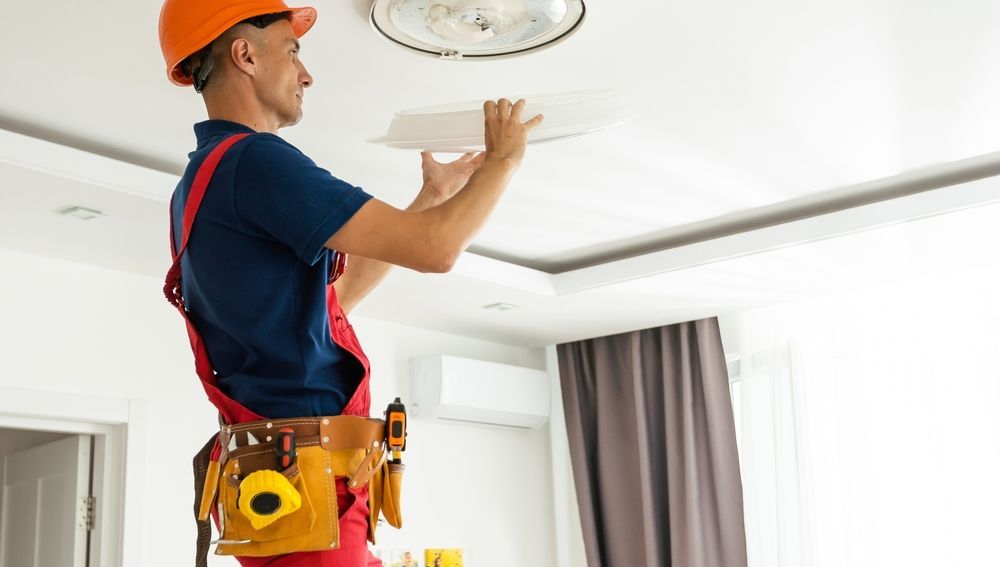 Man in hard hat and overalls installing ceiling panel near a light fixture.