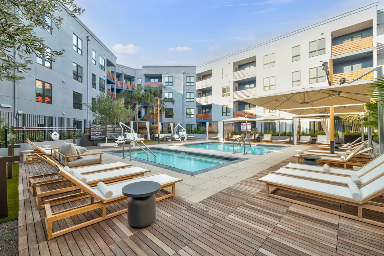 Pool area with lounge chairs, umbrellas, and a multi-story apartment building.